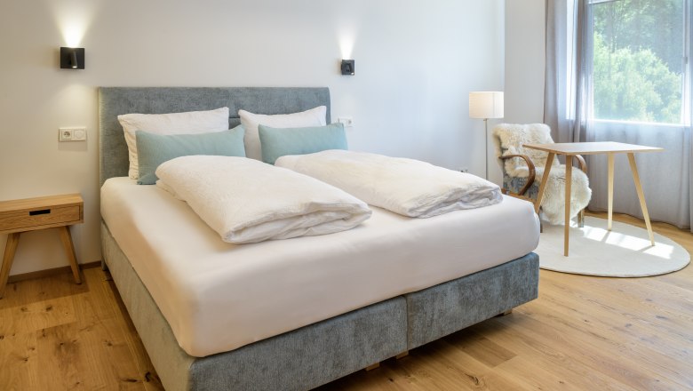 Ein modernes Schlafzimmer mit einem gro&szlig;en Bett, Holzboden, einem kleinen Tisch und einem Stuhl mit Fell&uuml;berwurf. Tageslicht str&ouml;mt durch ein Fenster.