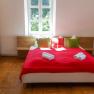 Schlafzimmer mit rotem Bett&uuml;berwurf, Kissen und Handt&uuml;chern auf dem Bett, Holzschrank und Fenster im Hintergrund.