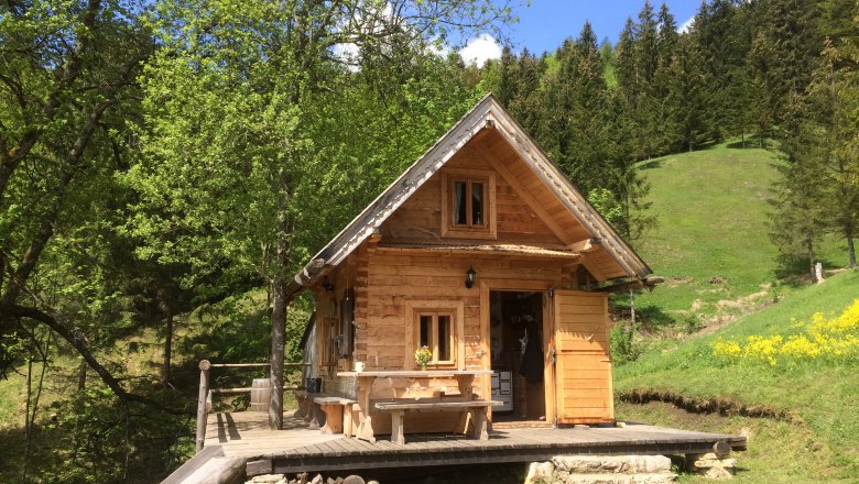 Eine kleine Holzhütte in einer grünen, bewaldeten Landschaft mit blauem Himmel.