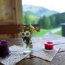 Ein Holztisch mit zwei Kerzen und einem kleinen Blumenstrau&szlig; in einem Glasgef&auml;&szlig;, im Hintergrund eine verschwommene Berglandschaft.