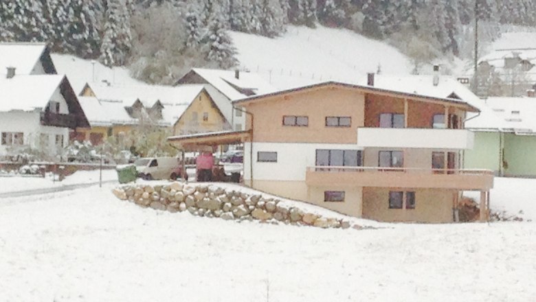 Modernes Haus im Schnee mit umliegenden Geb&auml;uden und Wald im Hintergrund.