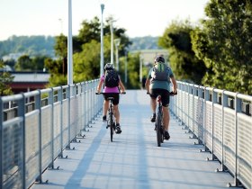 A couple cycle relaxed over a modern bridge, surrounded by lush greenery and the gentle beauty of nature. The warm rays of the sun bathe the scenery in a golden light and invite you to enjoy the fresh air and picturesque landscape.