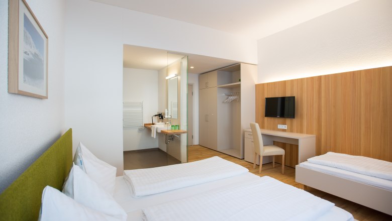 Modern furnished hotel room with two beds, desk, TV and open bathroom.