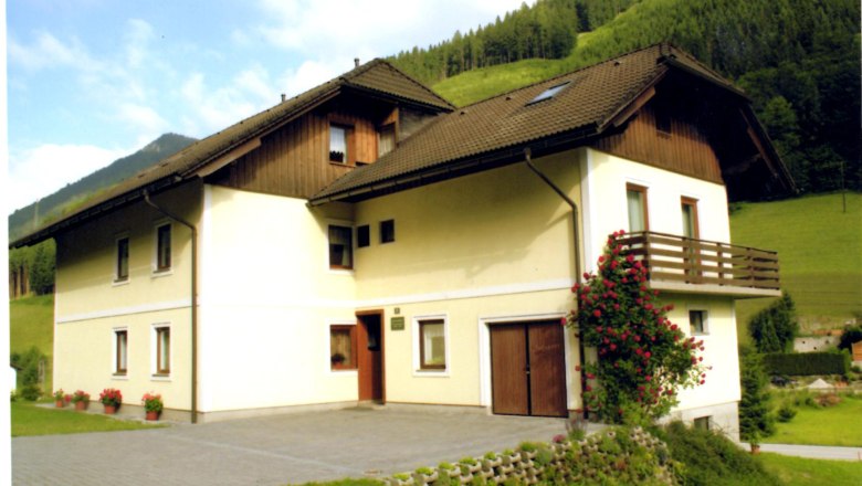 Ein zweistöckiges Haus mit Balkon und Rosenstrauch, umgeben von grünen Hügeln.