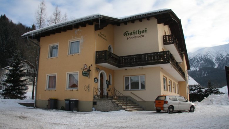 Family Pension Sonnenhof, © Ferenc Dudas A yellow building with the inscription 'Gasthof Sonnenhof' in the snow, surrounded by mountains.