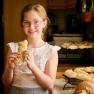 A girl with glasses holds a pastry in her hand and smiles at the camera. Other baked goods can be seen on a table in the background.