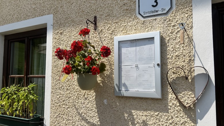 Au&szlig;enansicht einer Hauswand mit einer Adresse, einem Blumenarrangement und einem Herz aus Metall.