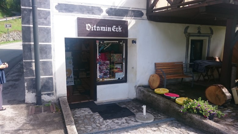 Eingang eines kleinen Ladens namens Vitamin-Eck mit Sitzbank und Dekorationen aus Holz und Blumen.