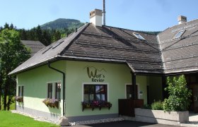 A green building with the inscription 'Wolf's Revier' and flower boxes in front of the windows, surrounded by trees and mountains in the background.