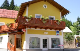 Yellow building with wooden veranda and flower boxes, hairdressing salon and savings bank on the first floor.