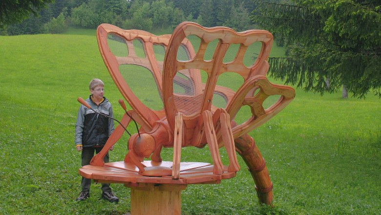 Ein Junge steht neben einer großen Holzskulptur eines Schmetterlings auf einer Wiese.