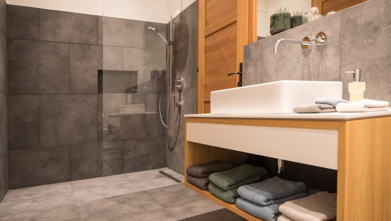 Modernes Badezimmer mit grauen Fliesen, Glasdusche und Holzwaschtisch.