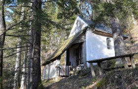 A small white chapel in the forest, surrounded by trees and a wooden table in the foreground.