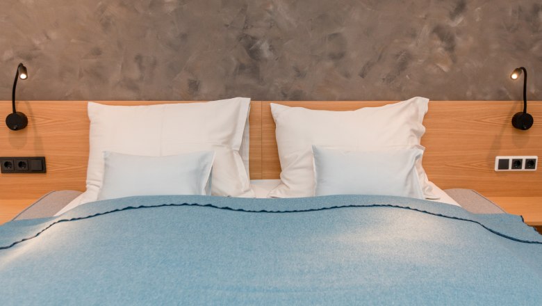 A double bed with a blue blanket and white pillows in front of a gray wall.
