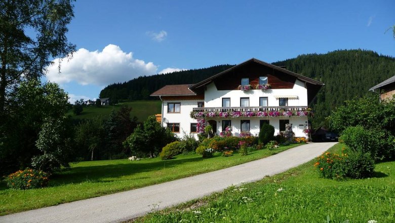 Ein Bauernhof mit Blumen geschmücktem Haus in einer grünen Landschaft unter blauem Himmel.