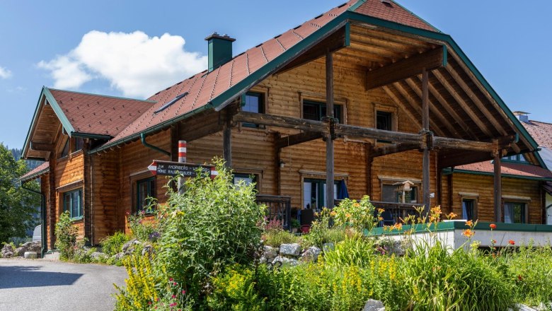 Biohof Herdengl in Lunz am See, © Theo Kust Ein traditionelles Holzhaus mit einem roten Dach, umgeben von bunten Blumen und grünen Pflanzen, unter einem blauen Himmel.