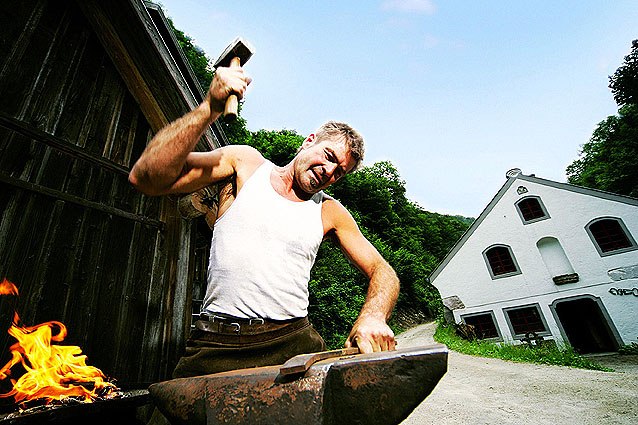 A blacksmith works with a hammer on an anvil in front of an outdoor building.