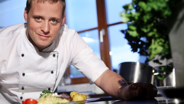 A chef in a white uniform presents a dish in a kitchen.