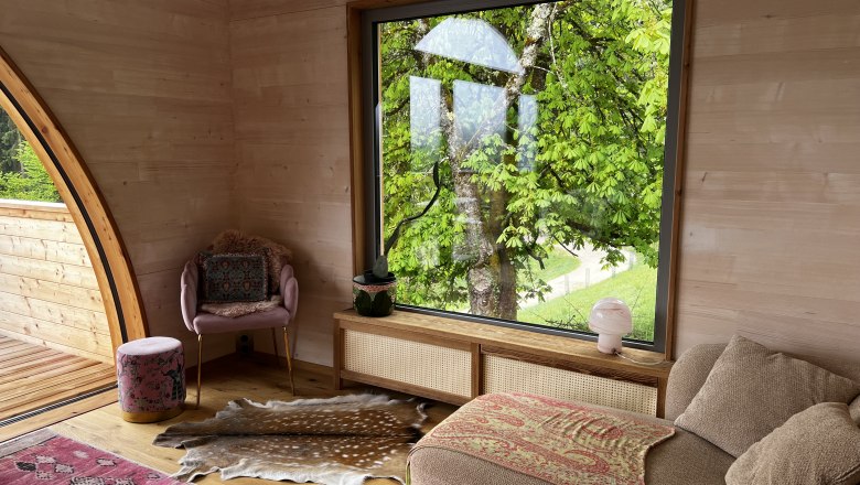 Innenansicht eines gem&uuml;tlichen Raumes mit gro&szlig;em Fenster, das auf einen gr&uuml;nen Baum blickt. Ein Sessel, ein Hocker und ein Sofa sind zu sehen.
