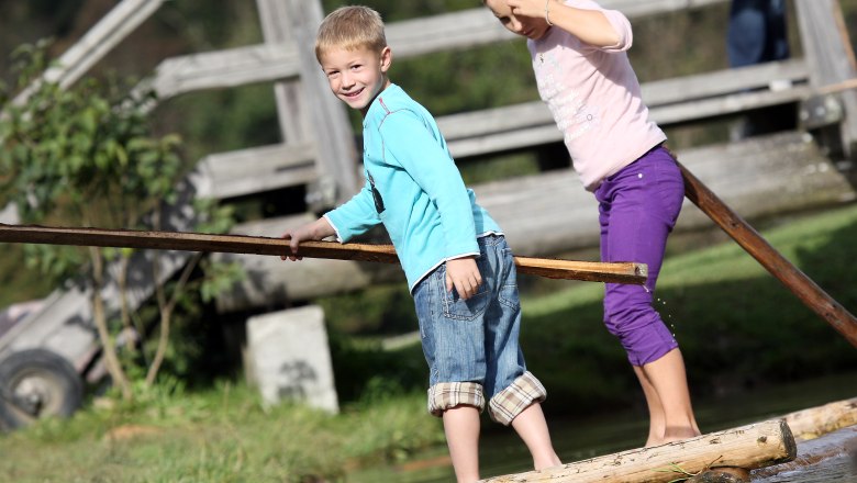 Two children balance on a wooden raft over water, in the background a wooden bridge.
