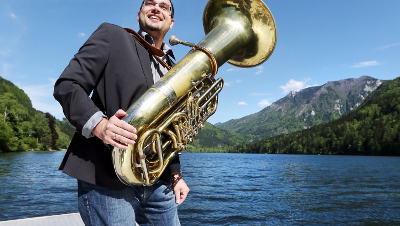 Ein Mann mit einer Tuba steht barfuß auf einem Steg an einem See, umgeben von Bergen und blauem Himmel.