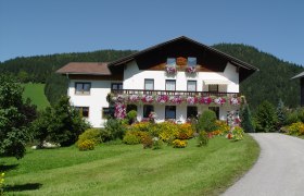 Ein wei&szlig;es Haus mit Blumenbalkonen vor einem bewaldeten H&uuml;gel.