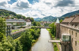 Panorama einer Flusslandschaft mit Schloss und moderner Architektur, umgeben von grünen Hügeln.