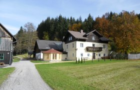 A large, two-story house with a balcony, surrounded by meadows and trees in autumn.