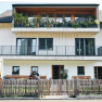 Ein modernes dreist&ouml;ckiges Haus mit Holzbalkonen und Pflanzen. Vor dem Haus sitzen zwei Personen auf einer Bank hinter einem Holzzaun.