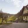 Ein gelbes Haus mit Balkon steht neben einem Spielplatz und einer Stra&szlig;e, die durch gr&uuml;ne H&uuml;gel f&uuml;hrt. Der Himmel ist klar und die Sonne scheint.