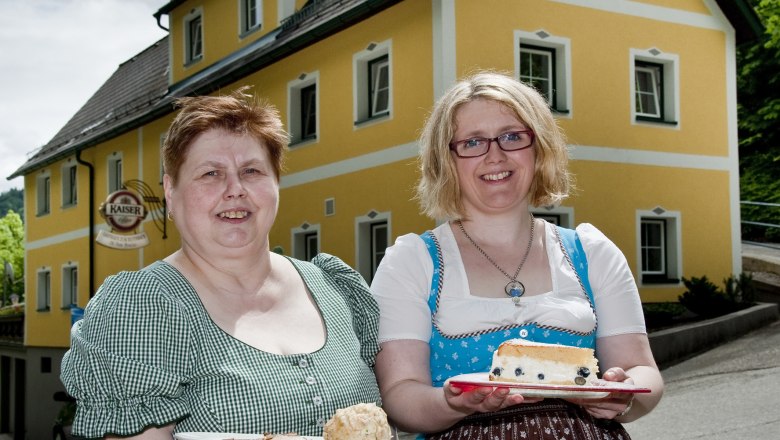 Zwei Frauen in traditioneller Kleidung halten Teller mit Speisen vor einem gelben Gebäude.