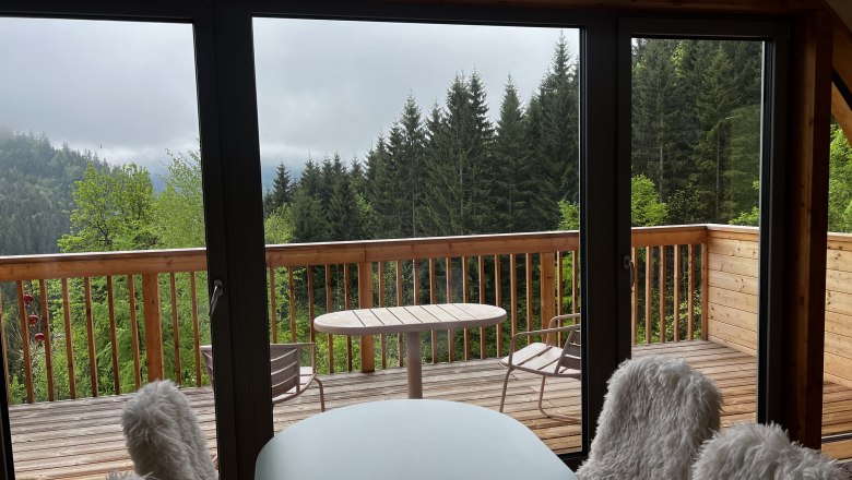 Blick von einem Innenraum auf eine Holzterrasse mit Wald im Hintergrund.
