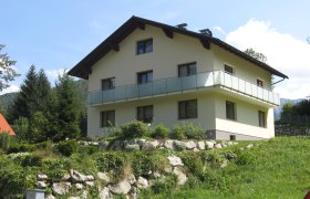 A two-storey house with balcony in a green, hilly landscape.