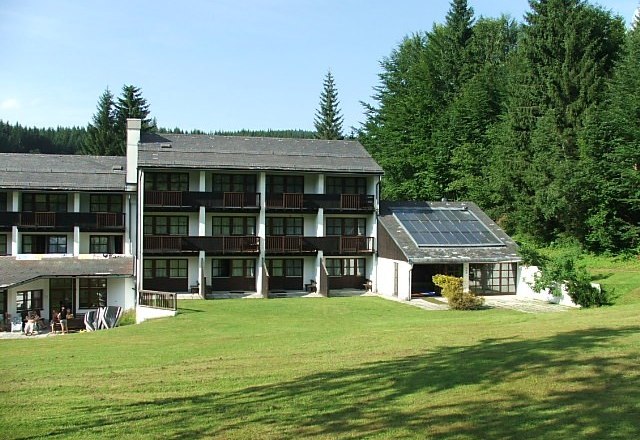 Hotel building with solar panels and green meadow in the foreground, surrounded by forest.