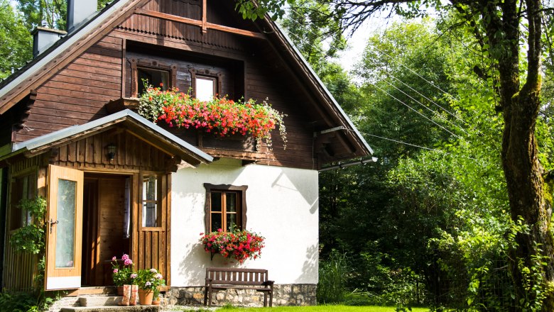 Ein malerisches Ferienhaus mit Holzverkleidung und Blumenk&auml;sten, umgeben von &uuml;ppigem Gr&uuml;n.