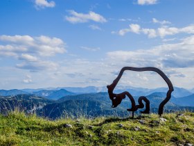 Ein majest&auml;tischer Blick auf die sanften H&uuml;gel und die beeindruckenden Berge, die sich unter einem strahlend blauen Himmel erstrecken. Die harmonische Verbindung von Natur und Kunst wird durch die stilvolle Skulptur des &Ouml;tscherb&auml;ren unterstrichen, die den Wanderern ein Gef&uuml;hl von Abenteuer und Freiheit vermittelt.