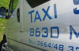 Nahaufnahme eines silbernen Taxis mit blauer Aufschrift und Telefonnummer.