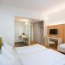 Modern furnished hotel room with two beds, desk, TV and open bathroom.
