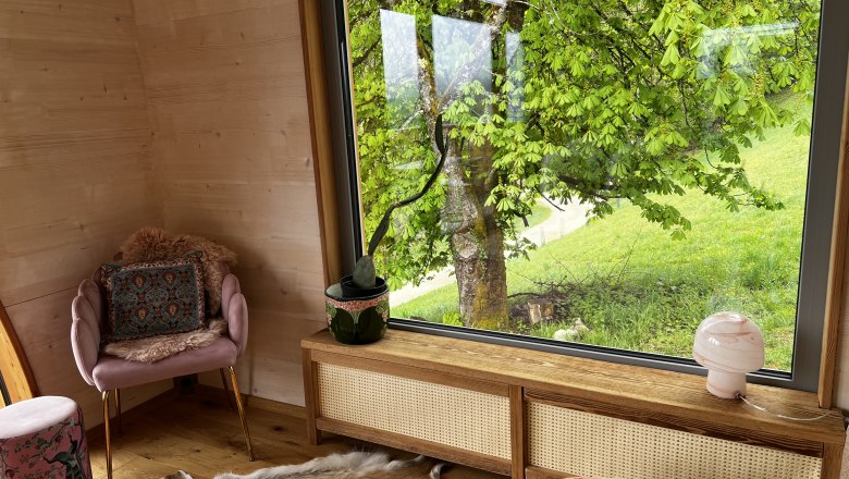 Innenraum eines gem&uuml;tlichen Zimmers mit gro&szlig;em Fenster und Blick auf einen Baum und gr&uuml;ne Wiese.