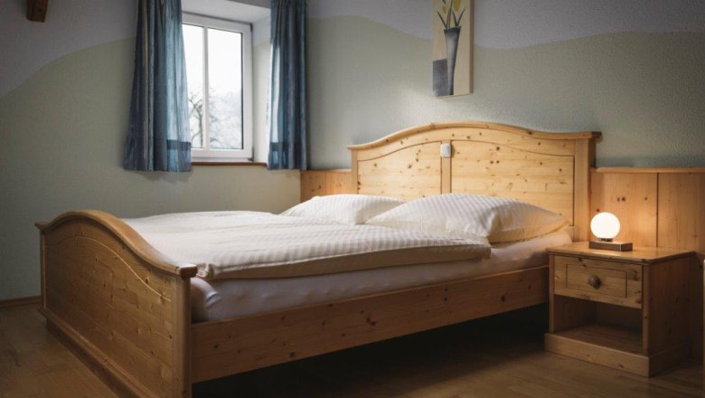 Ein Schlafzimmer mit einem Holzbett, Nachttisch und Lampe, blauen Vorh&auml;ngen und einem Bild an der Wand.