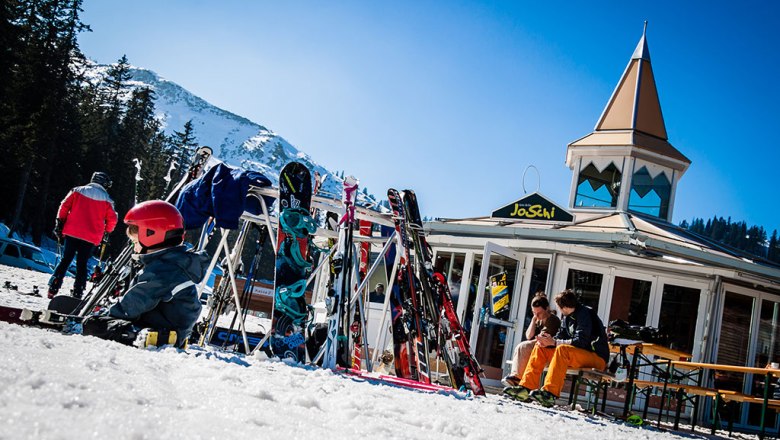 Skiers in front of the JoSchi bar with skis and snowboards in the snow.
