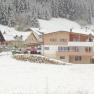 Modern house in the snow with surrounding buildings and forest in the background.