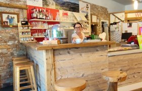 Eine Frau steht hinter einer rustikalen Holztheke in einer gem&uuml;tlichen Lounge mit Holzhockern und Steinwand.