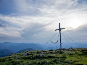 Ein majest&auml;tisches Gipfelkreuz thront &uuml;ber den sanften H&uuml;geln, umgeben von einer atemberaubenden Berglandschaft. Die Wolken ziehen sanft &uuml;ber den Himmel und verleihen der Szenerie eine friedliche Atmosph&auml;re, w&auml;hrend die frische Bergluft zum Wandern einl&auml;dt.