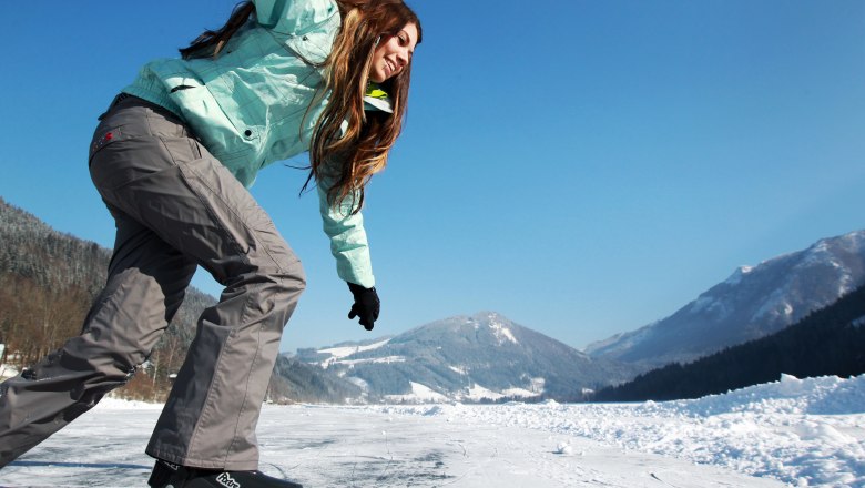 A person skates on a frozen lake with snow-covered mountains in the background.