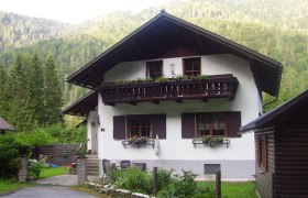 A traditional vacation home with a wooden balcony and flower boxes, surrounded by green trees and mountains.