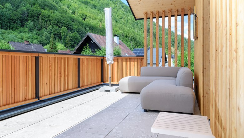 Moderne Terrasse mit Holzw&auml;nden, grauem Sofa und Blick auf bewaldete H&uuml;gel.