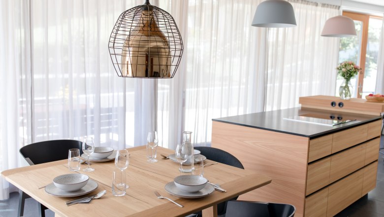 Modern eingerichtete K&uuml;che mit Holztisch, gedeckt f&uuml;r vier Personen, und K&uuml;cheninsel im Hintergrund.