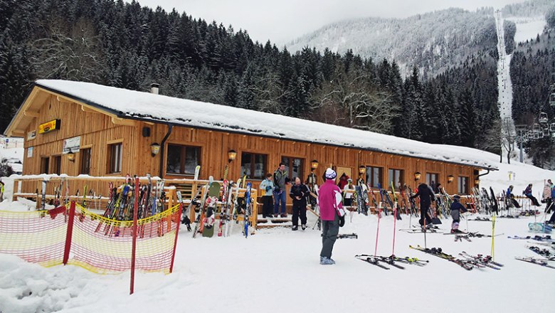 Ski hut in the snow with skiers and equipment in front of it.