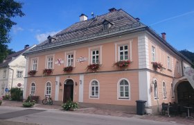 Ein zweistöckiges, rosa Gebäude mit weißen Fensterrahmen und Blumenkästen, beschriftet mit 'Rathaus'.